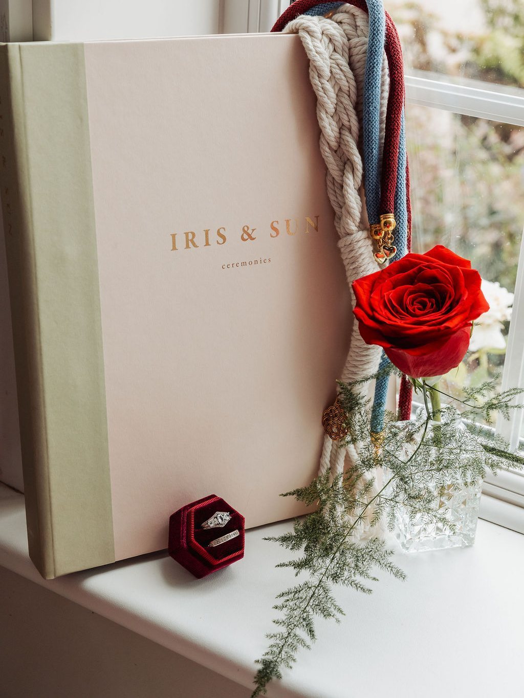 Jan's Iris and Sun ceremony folder on a windowsill with handfasting cords, a red rose and a ring box
