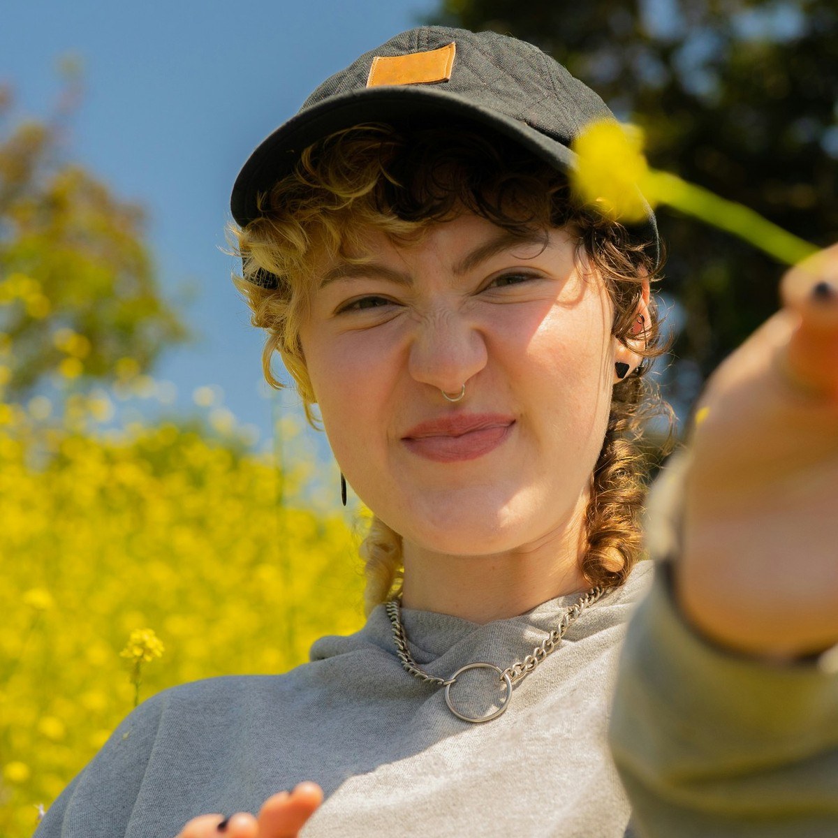 A smiling young person in a cap holds a yellow wildflower out in a sunny field